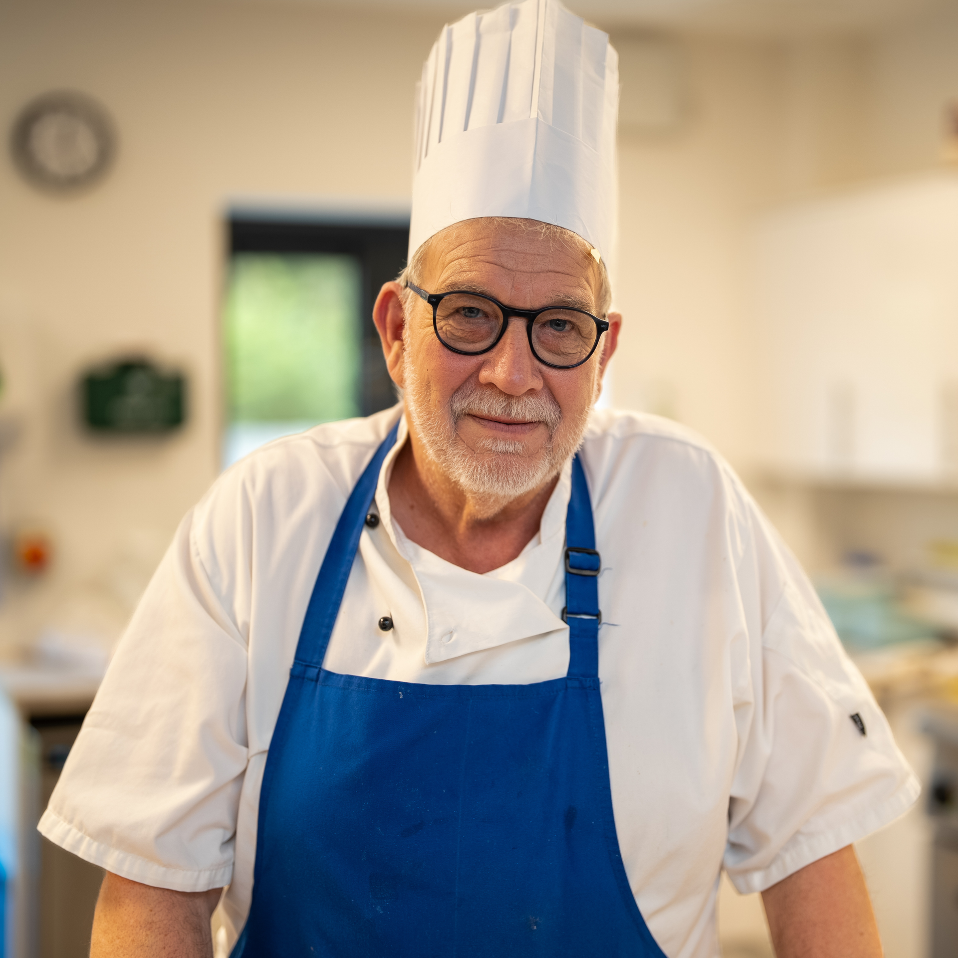 Chef smiling, wearing white chef's hat and coat, against a light blue background.