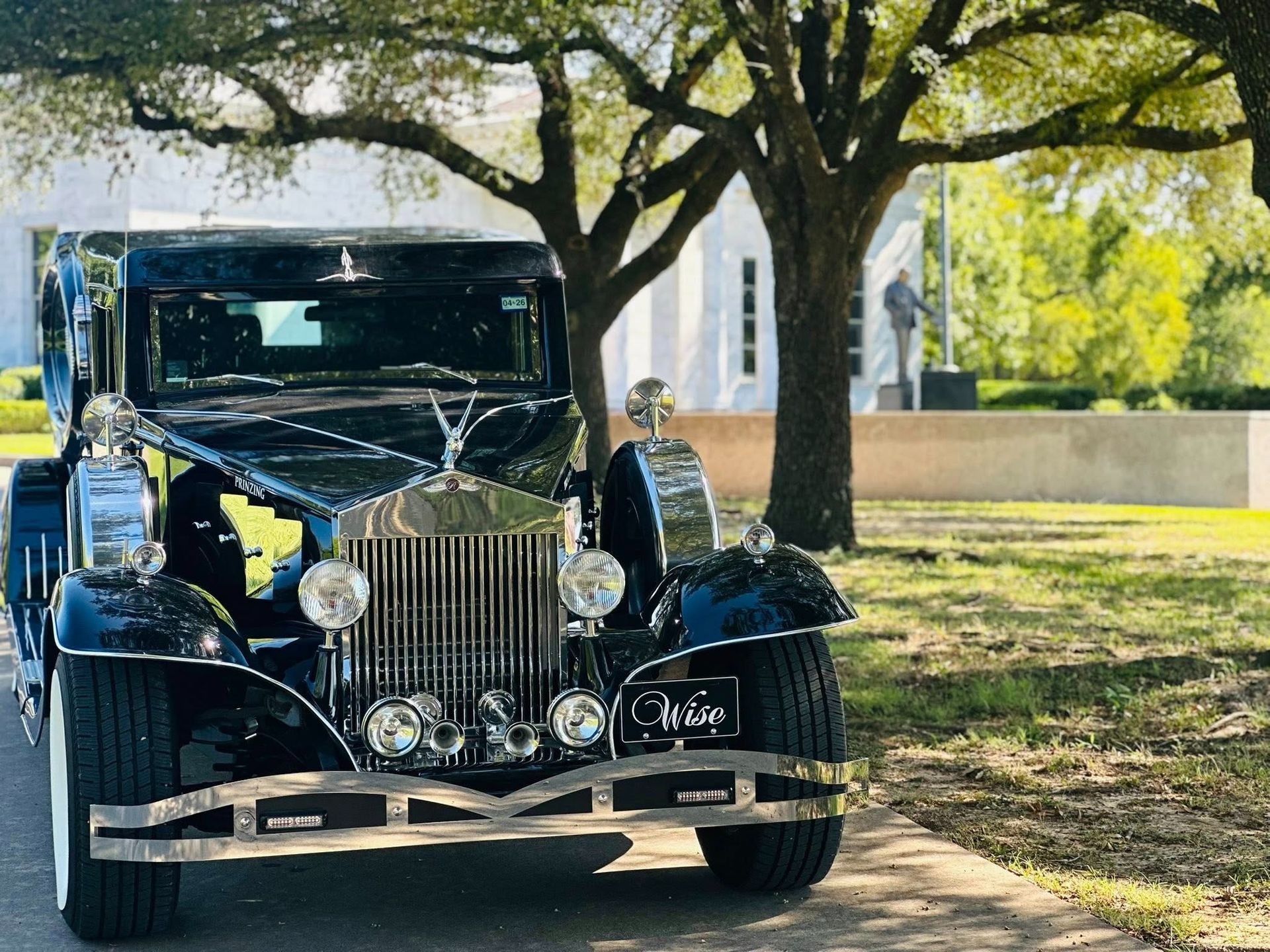 Black vintage car with chrome details, parked on a path with a building and large tree in the background.