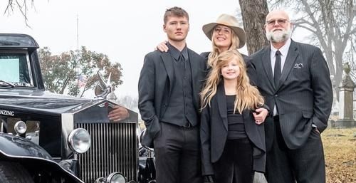 Family in formal attire posing next to a vintage black car, possibly a hearse, outdoors.