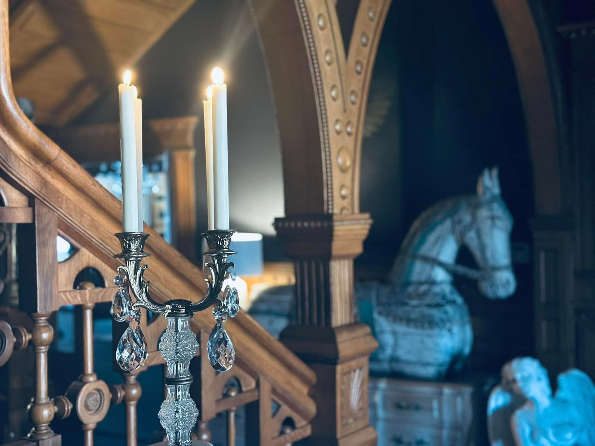 Candlelit chandelier on wooden staircase, ornate details. Horse statue in background.