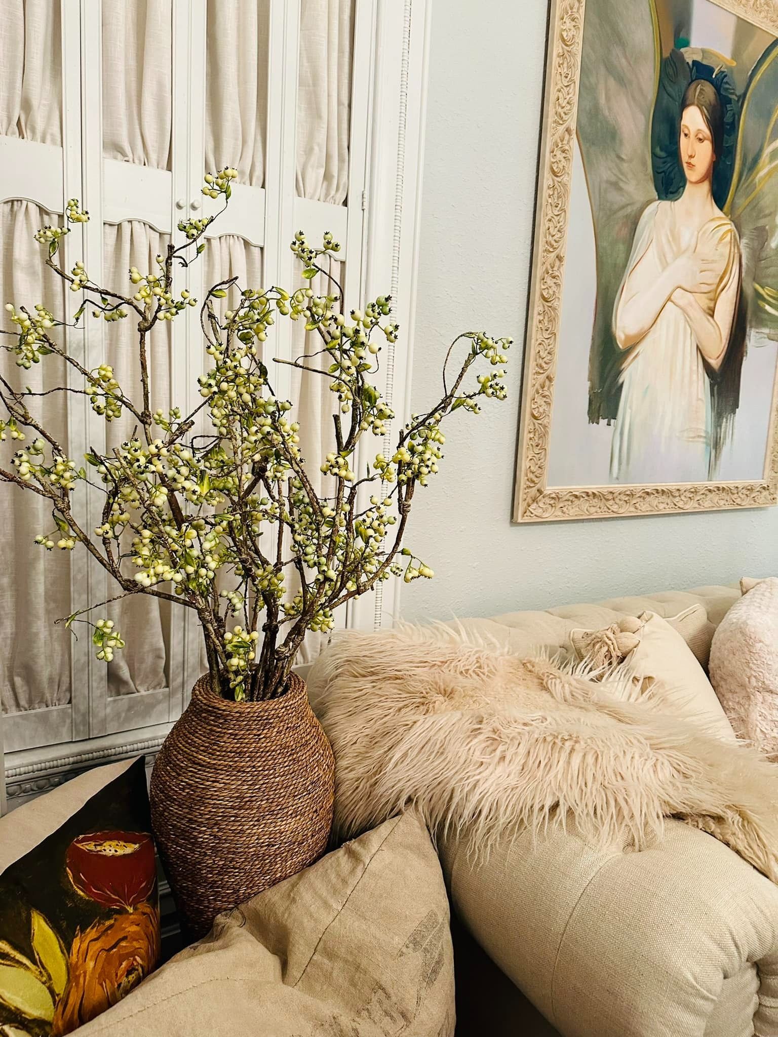 Vase of green branches on a beige couch with fluffy pillows, next to a framed artwork and sheer curtains.