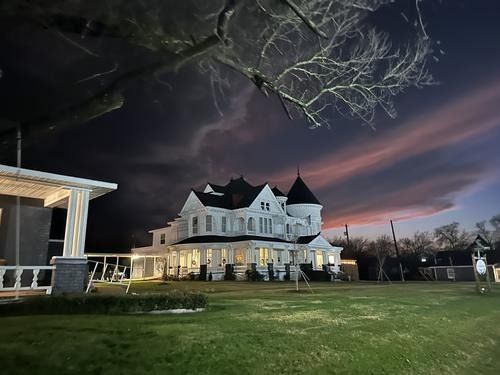 A large, white Victorian house under a dusky, colorful sky. Bare tree branches frame the top of the photo.