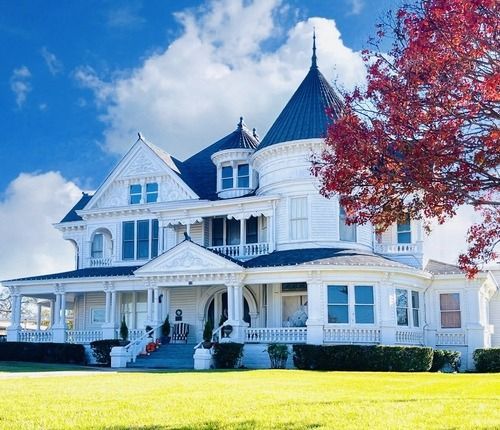 White Victorian house with a conical tower and wraparound porch against a blue sky.