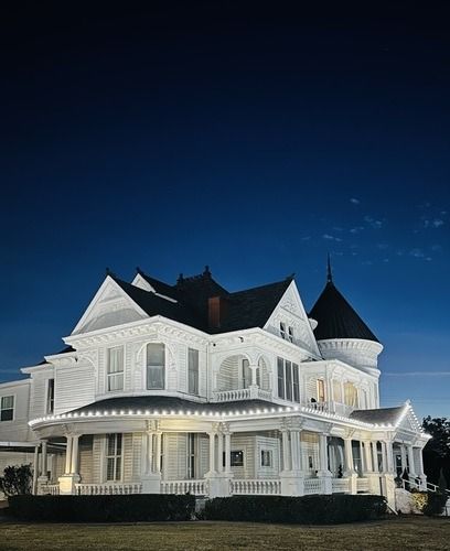 White Victorian house with porch, lit up at night, against a dark blue sky.