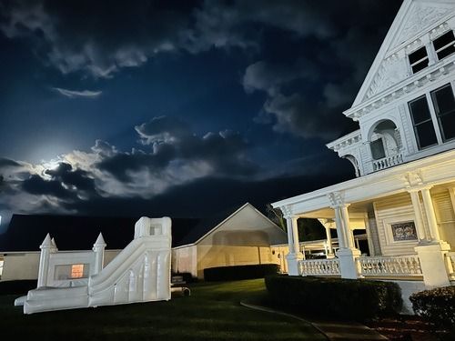 A white Victorian house illuminated at night with a cloudy sky and inflatable slide on the lawn.