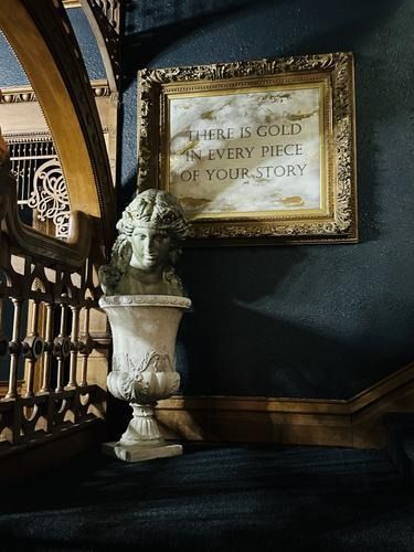 Gold-framed sign over stone bust, staircase backdrop; inscription: “There is gold every piece of your story.”