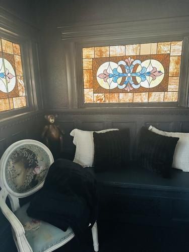 Cozy nook with stained-glass window, built-in bench with pillows, decorative chair, and teddy bear. Dark walls.