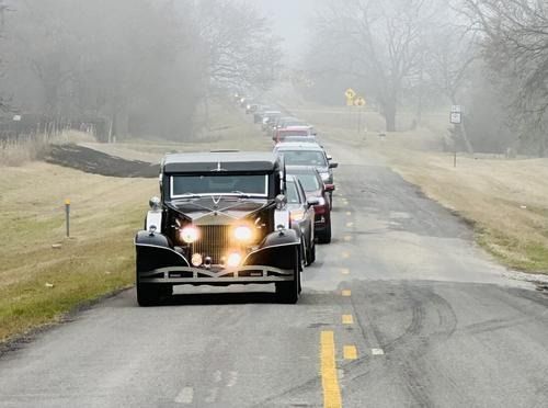 Line of cars, led by a vintage black car with headlights on, driving down a foggy road.