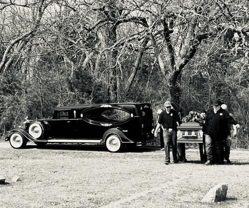 Funeral scene: Hearse, casket on a wheeled cart, men standing near, in a grassy cemetery. Black and white.
