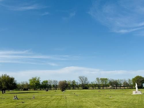 Open green field with scattered headstones, trees, and statue under a bright blue sky with wispy clouds.