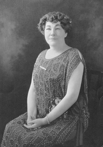 Woman in beaded dress, seated with hands clasped. Formal portrait, dark background.