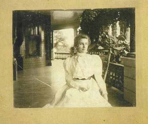 Woman in white dress sits on a porch, vines and railing in the background.
