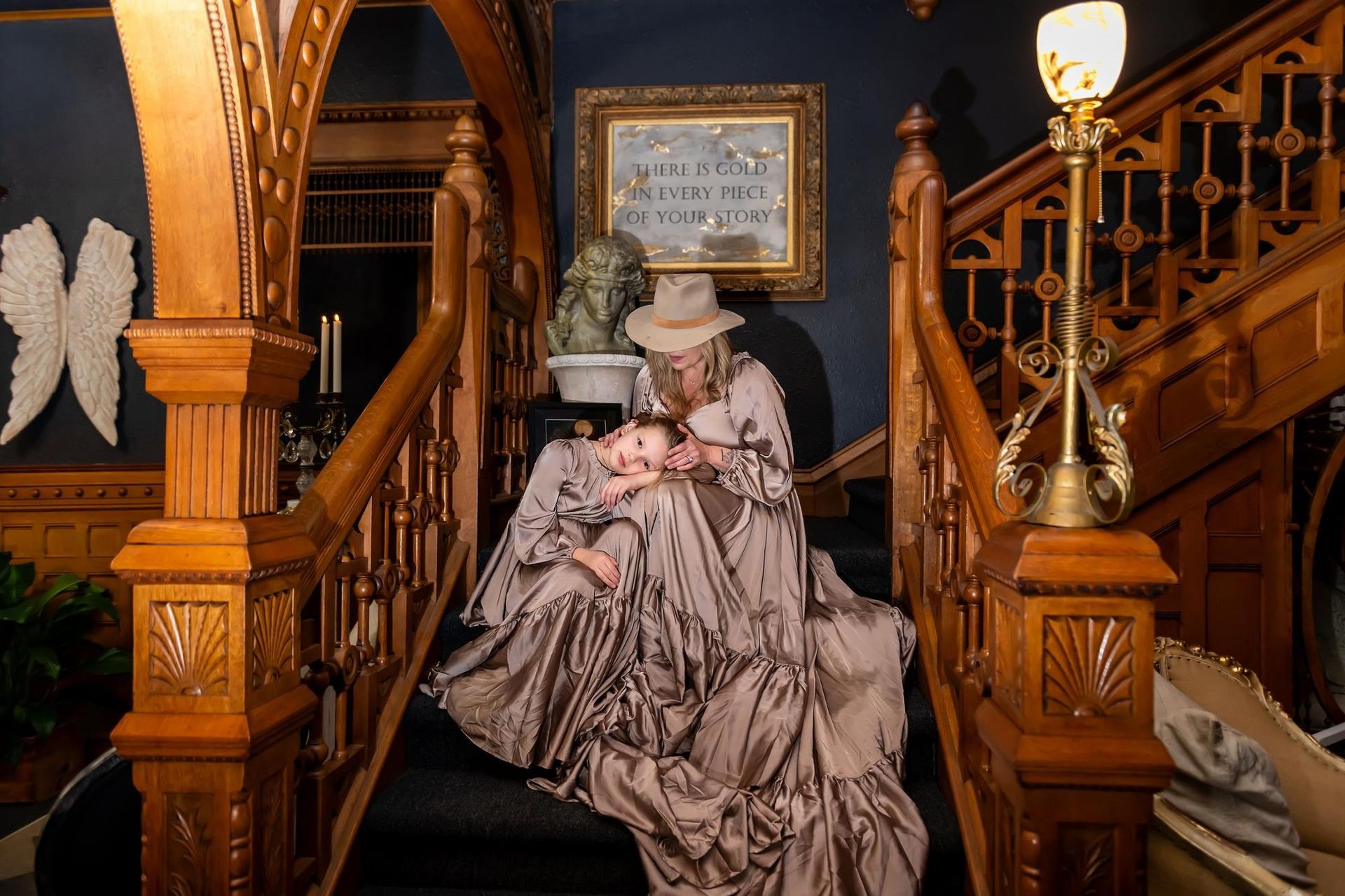 Woman in ornate dress on staircase holds baby. Brown wood, dark blue wall, framed art, and decorative light.