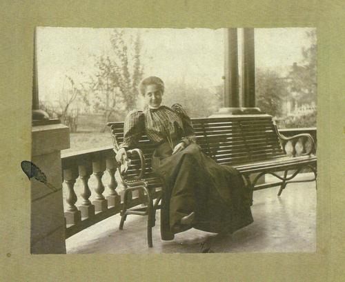 Woman in dark dress seated on bench on a porch.