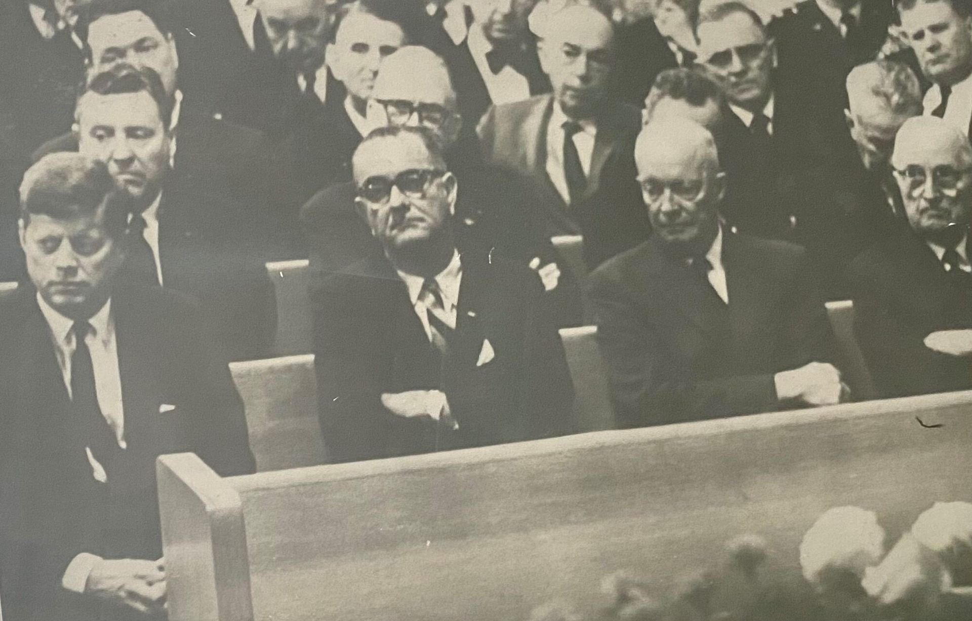 John F. Kennedy, Lyndon B. Johnson, and others seated during a somber event. Men in suits, black and white.