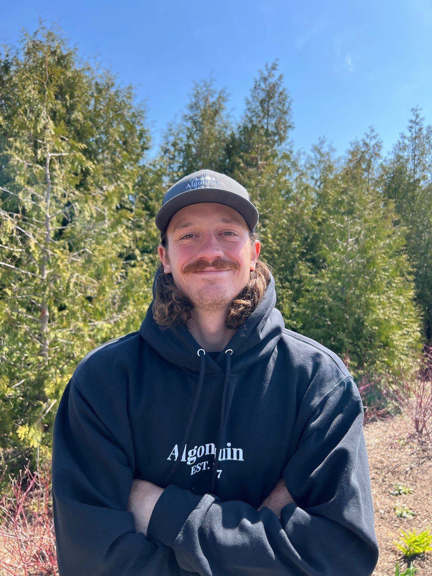 A man wearing a hat and a hoodie is standing in front of a forest.