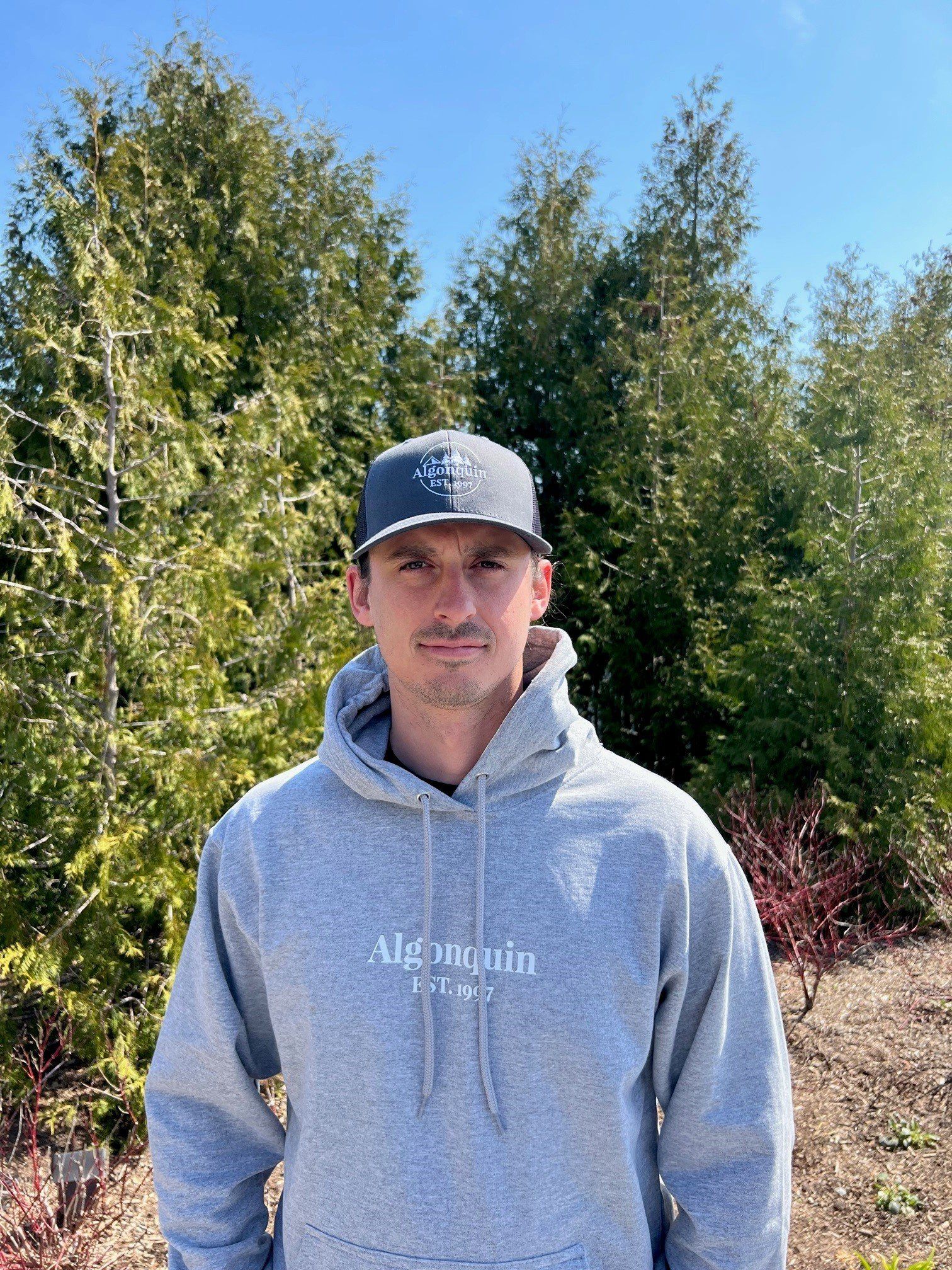 A man wearing a hoodie and a hat is standing in front of trees.