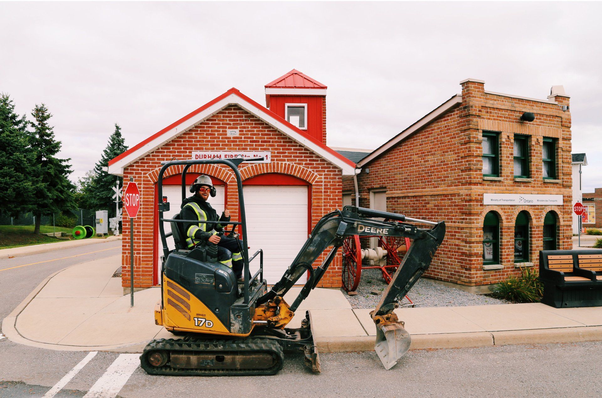 A man is driving a small excavator in front of a fire station.