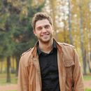 A man wearing a brown jacket and a black shirt is smiling