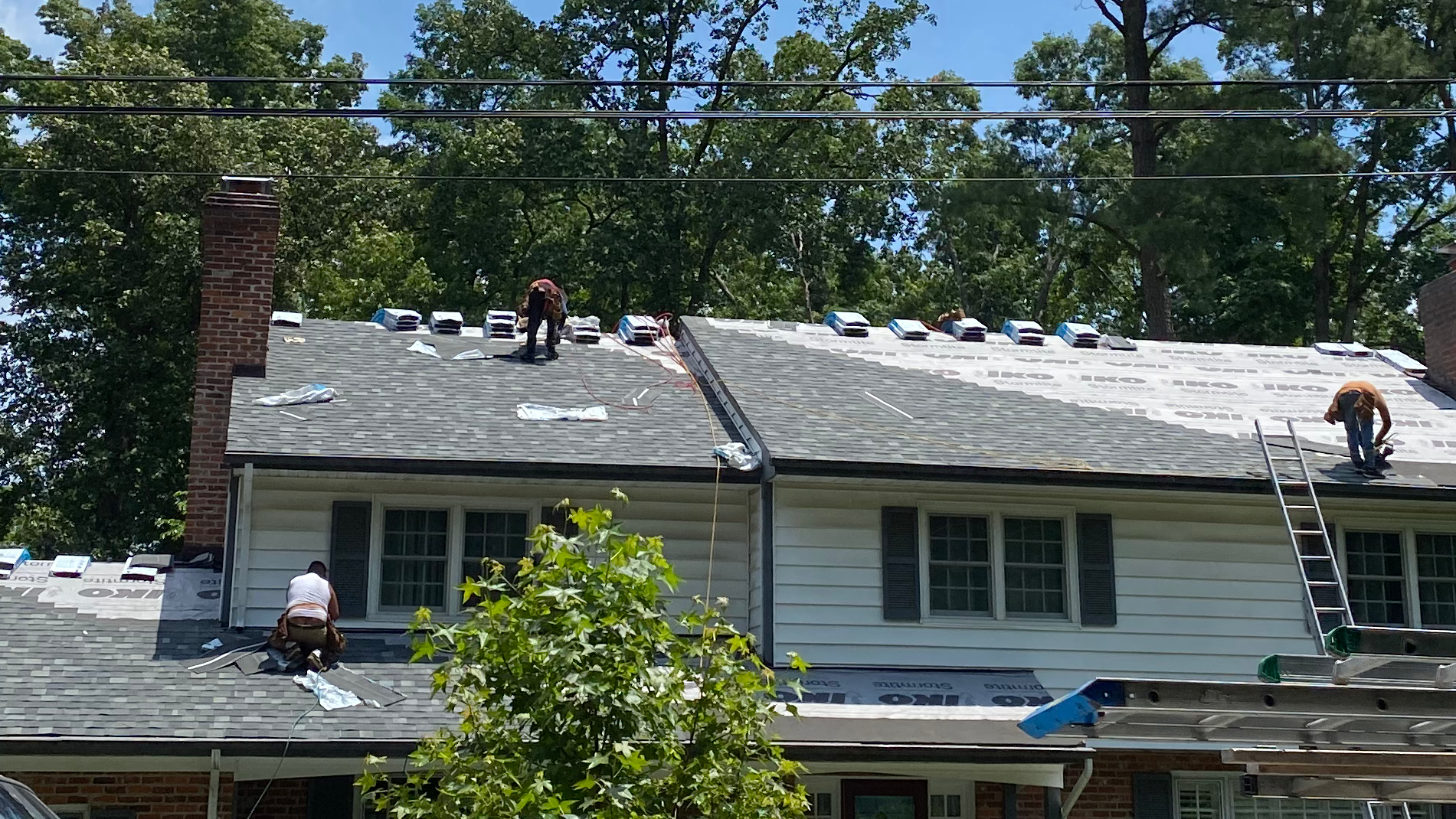 Workers on a roof repairing shingles on a house with a chimney and trees in the background