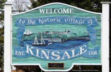 Welcome sign for the historic village of Kinsale, established in 1706. Features a coastal scene with sailing ships and trees, conveying a quaint, timeless feel.