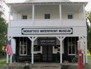 A white, two-story Morattico Waterfront Museum building with a porch, American flag, and vintage red gas pump.