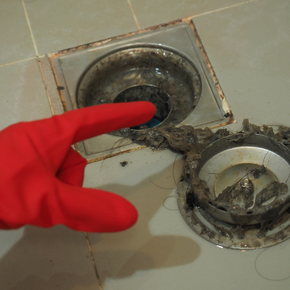 A person wearing a red rubber glove is pointing at a dirty drain