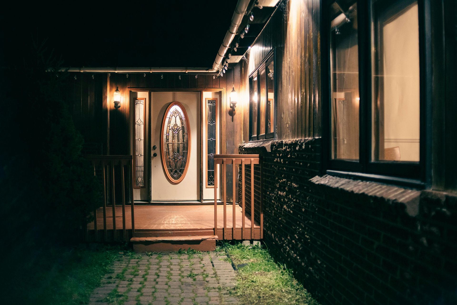 A house at night with porch lit by lights; front door with decorative glass, brown siding and trim, brick path.