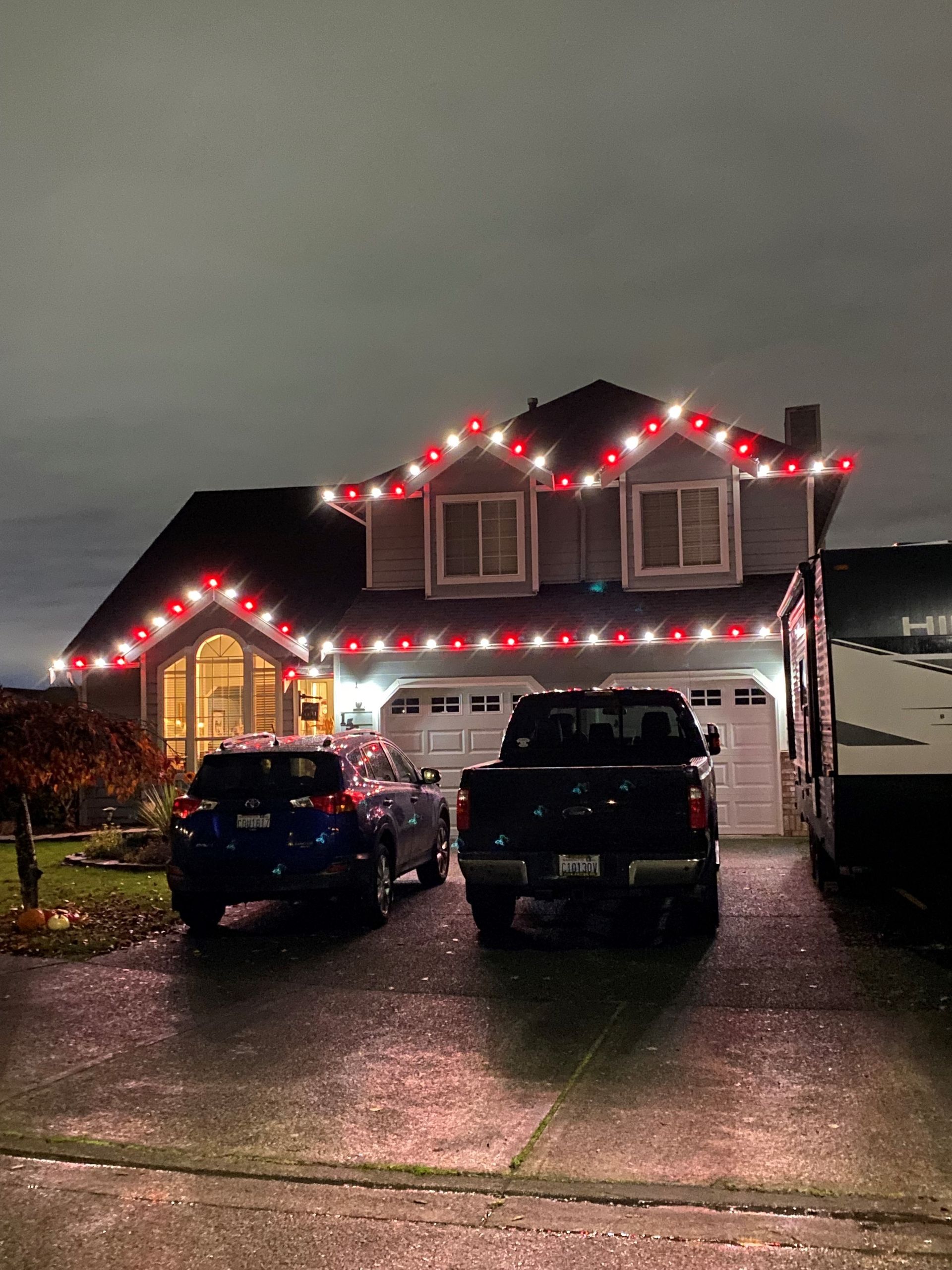 Two cars are parked in front of a house decorated with christmas lights.