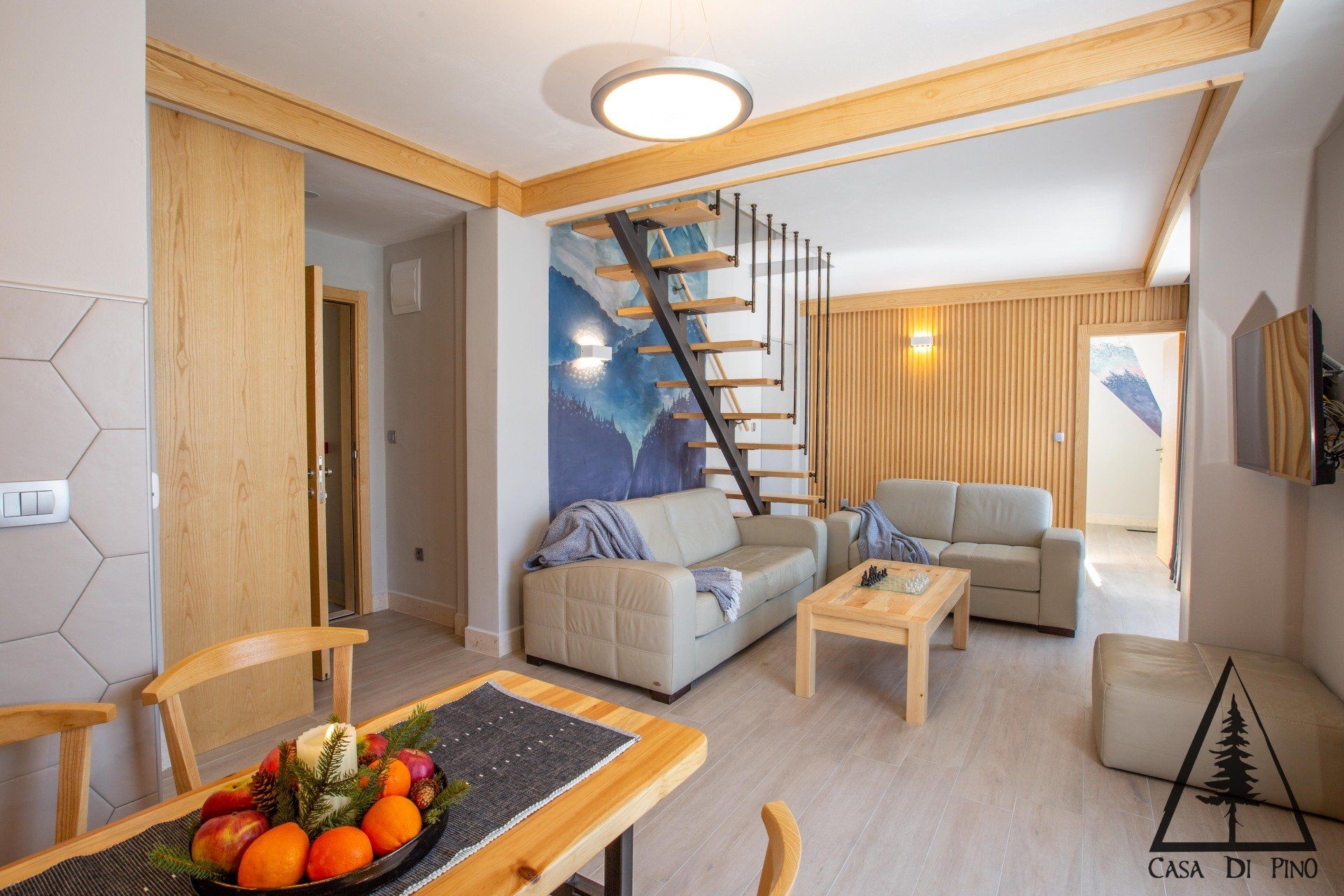 A living room with a big wall mural a couch, table, chairs and stairs leading up to the second floor.