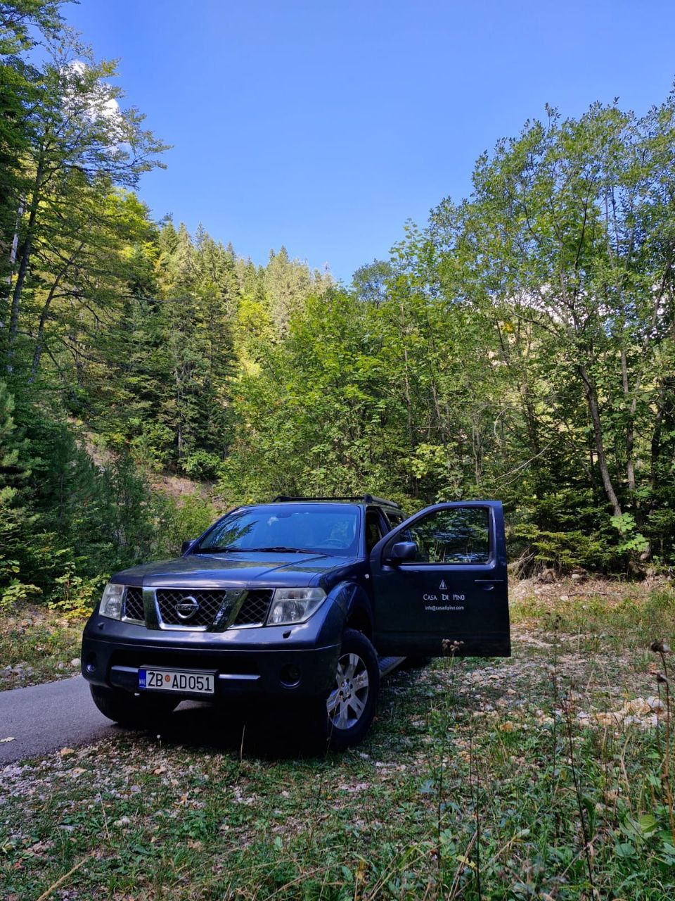 Dark blue Nissan Pathfinder parked on a forest path with an open door; trees and blue sky in the background.