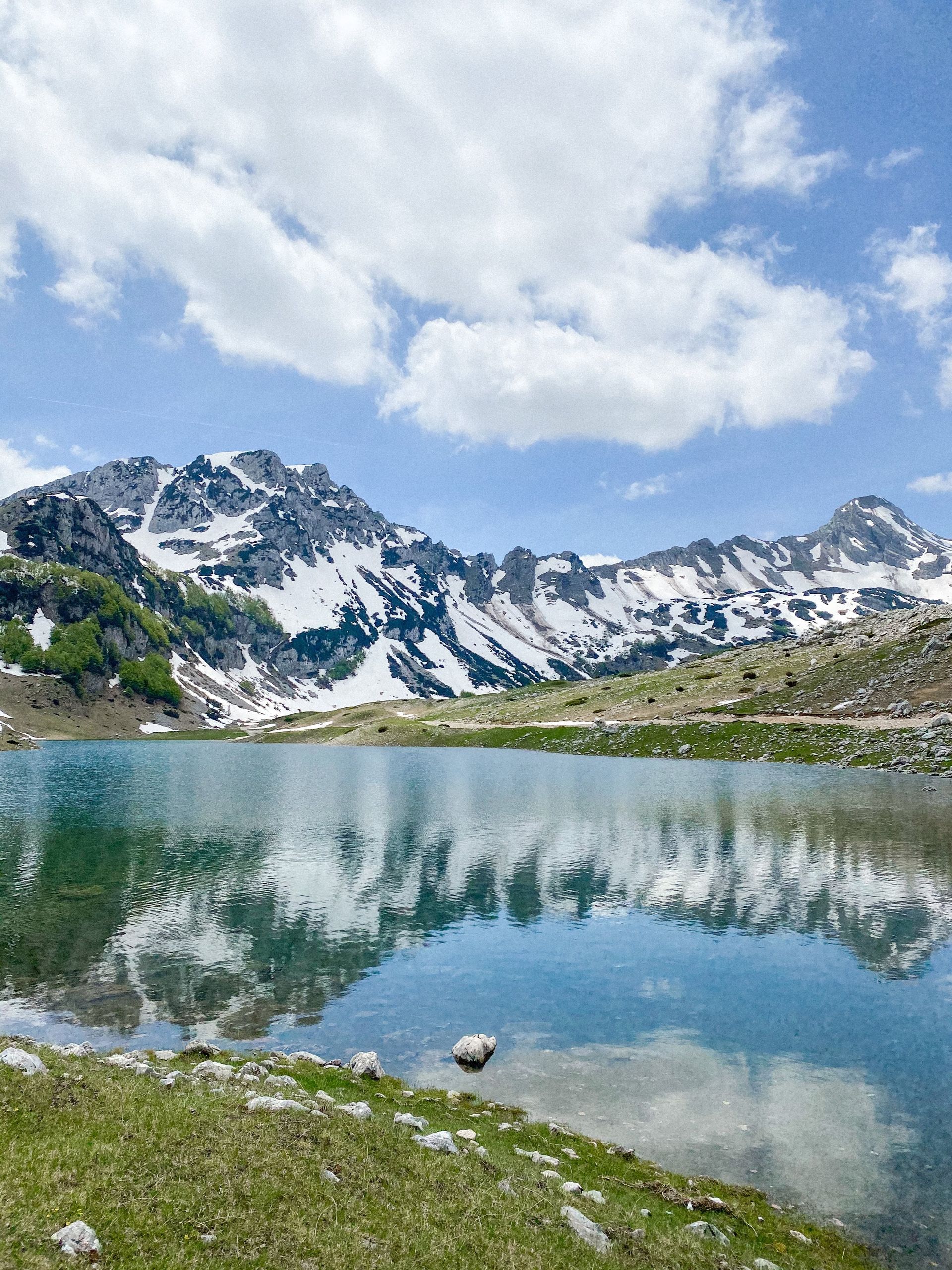Snow-capped mountains reflect in a still, turquoise lake under a partly cloudy sky. Green grass in foreground.