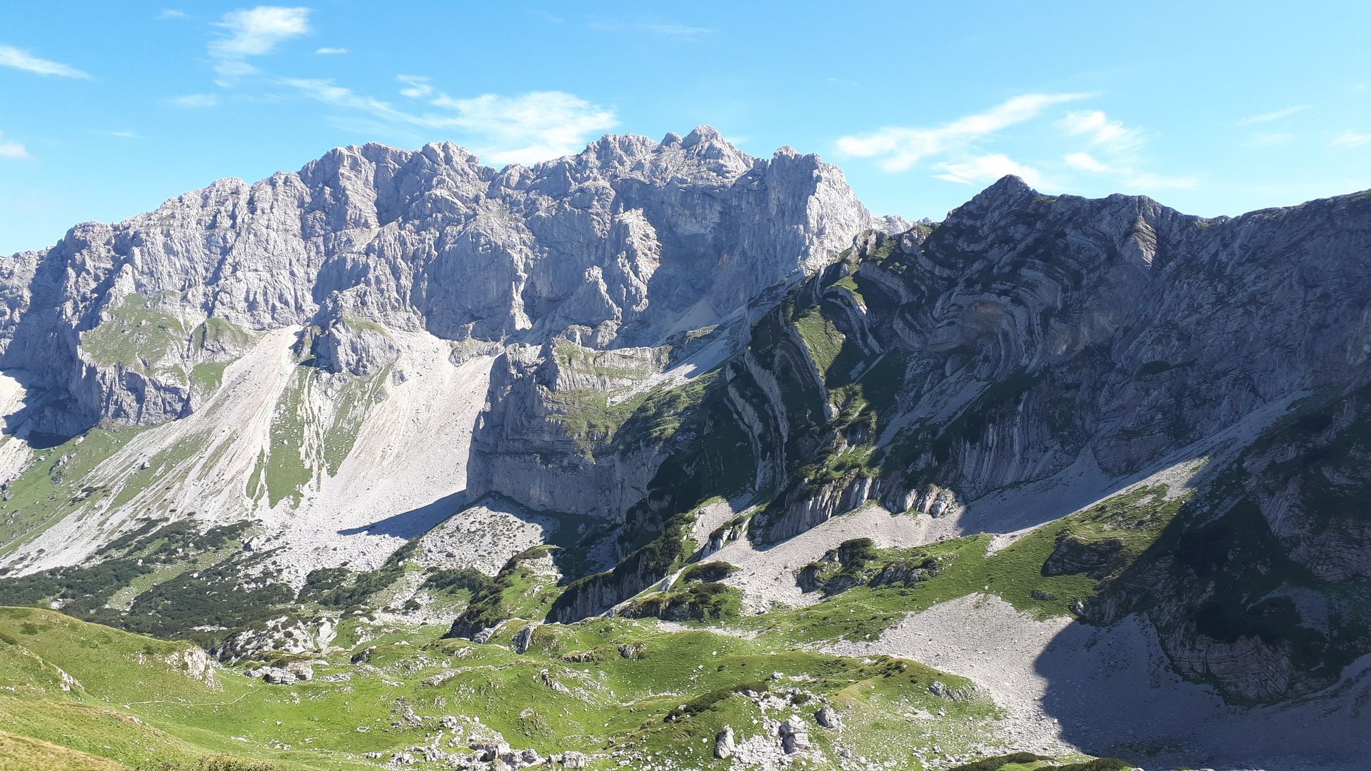 Rugged mountain range under a blue sky. Patches of green grass in the foreground, rocky terrain.