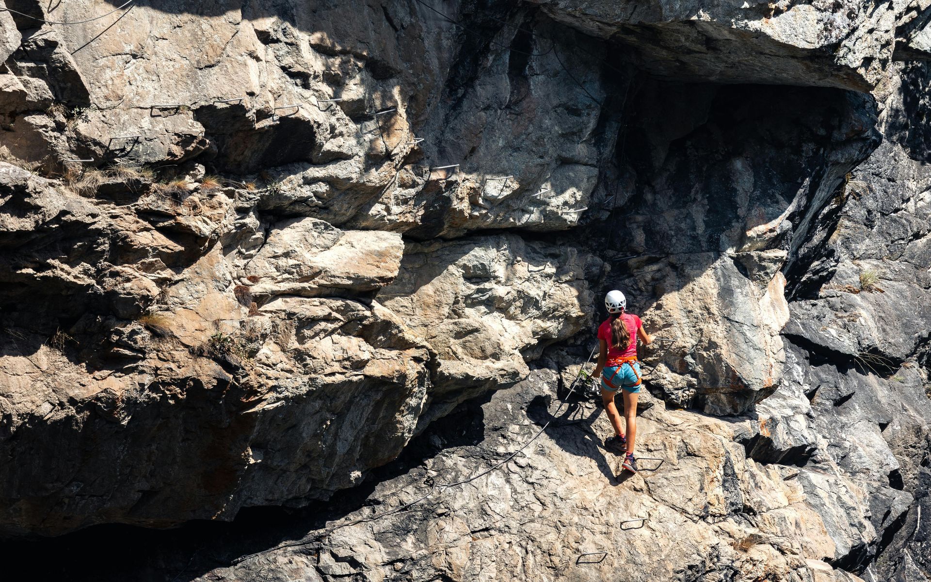 Rock climber ascends a sunlit, rugged rock face; wearing a red shirt, helmet, and blue shorts.