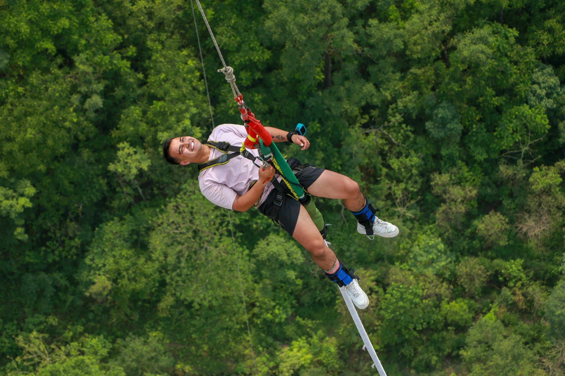 Man bungee jumping off a bridge into a green forest. He is wearing a harness and smiling.