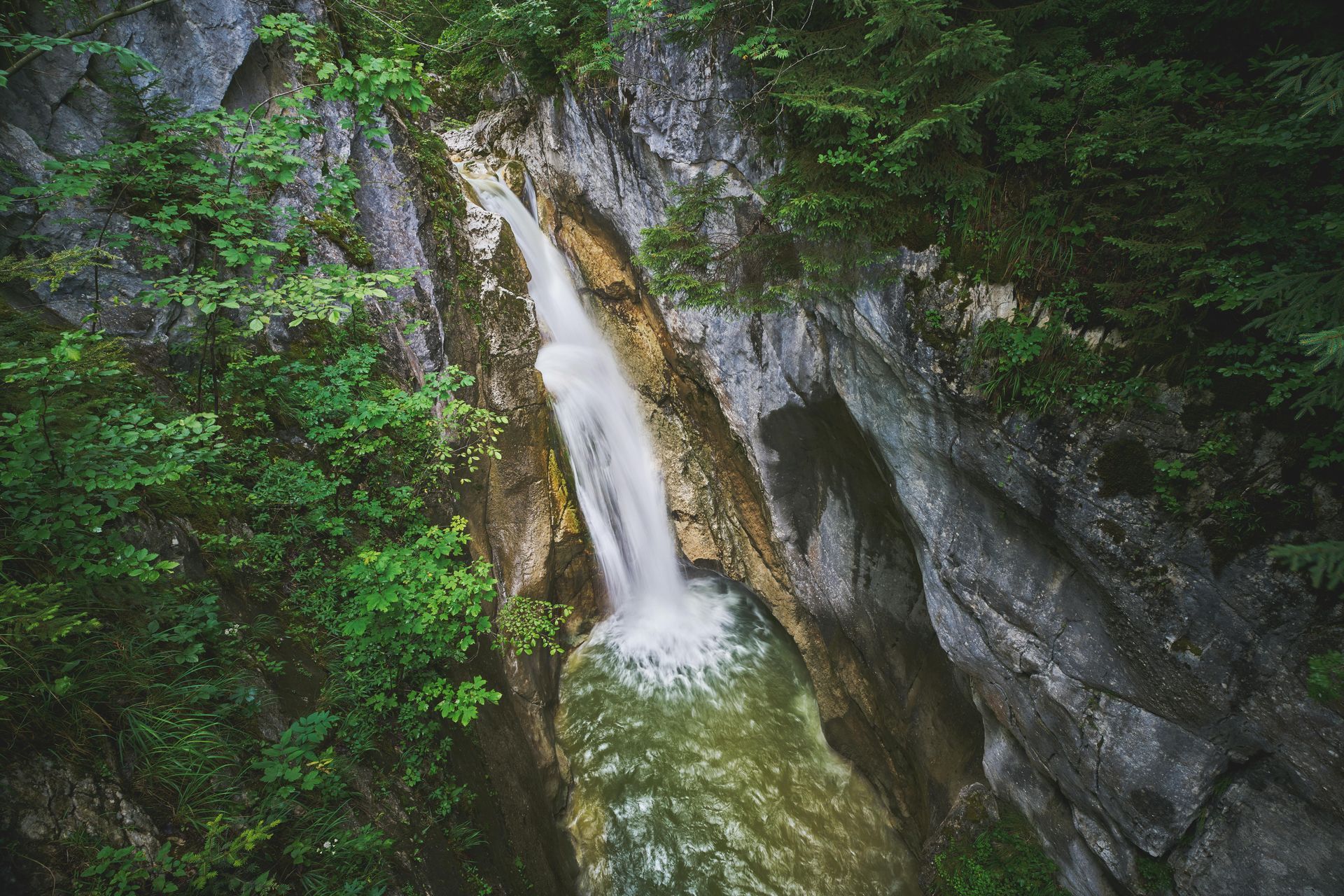 Waterfall cascading down a rocky canyon, surrounded by lush green trees.
