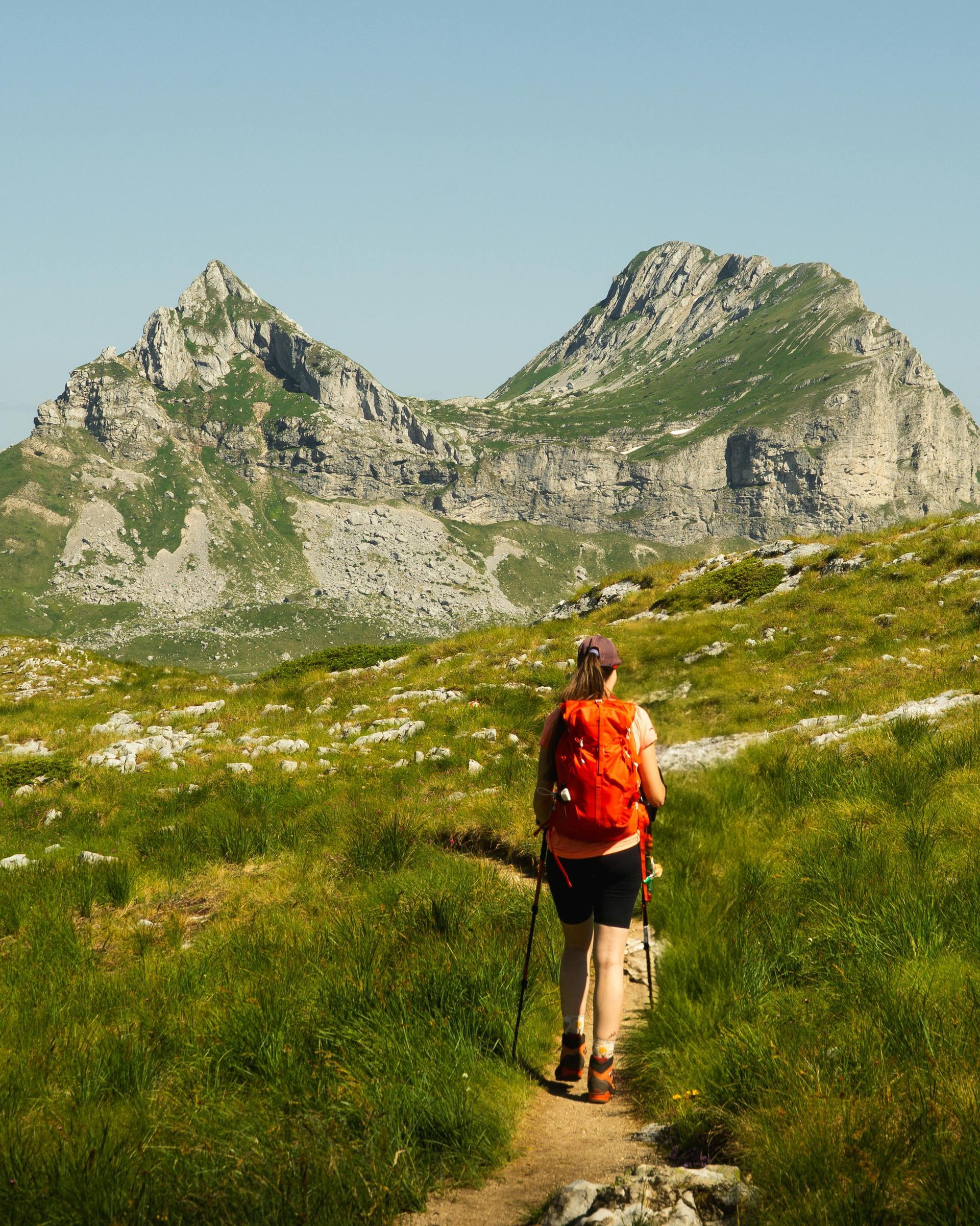 Woman hiking on a path towards two mountain peaks, wearing an orange backpack, green grass, blue sky.