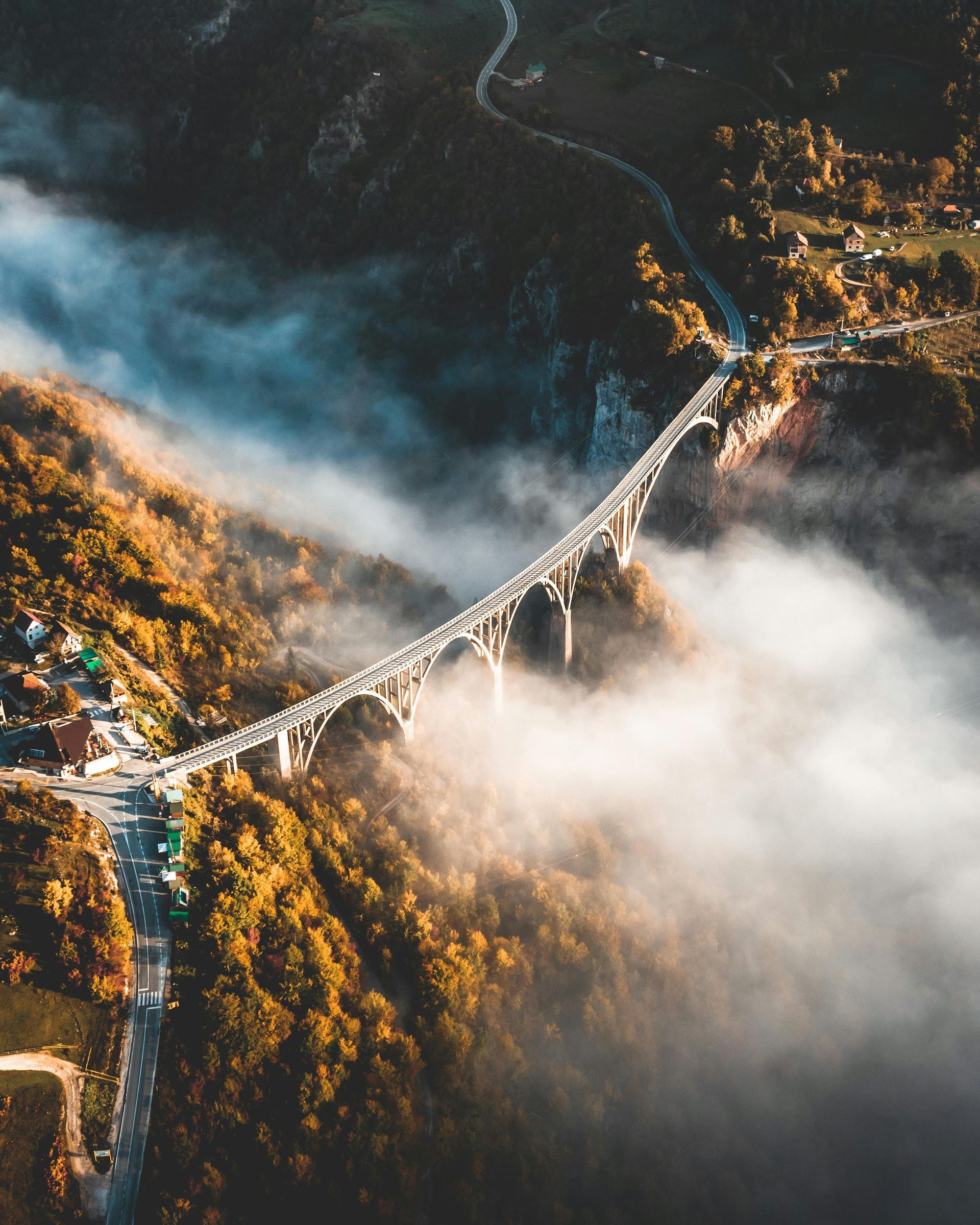 Bridge spanning a deep gorge, shrouded in mist. Autumn trees line the sides, road leads to the bridge.