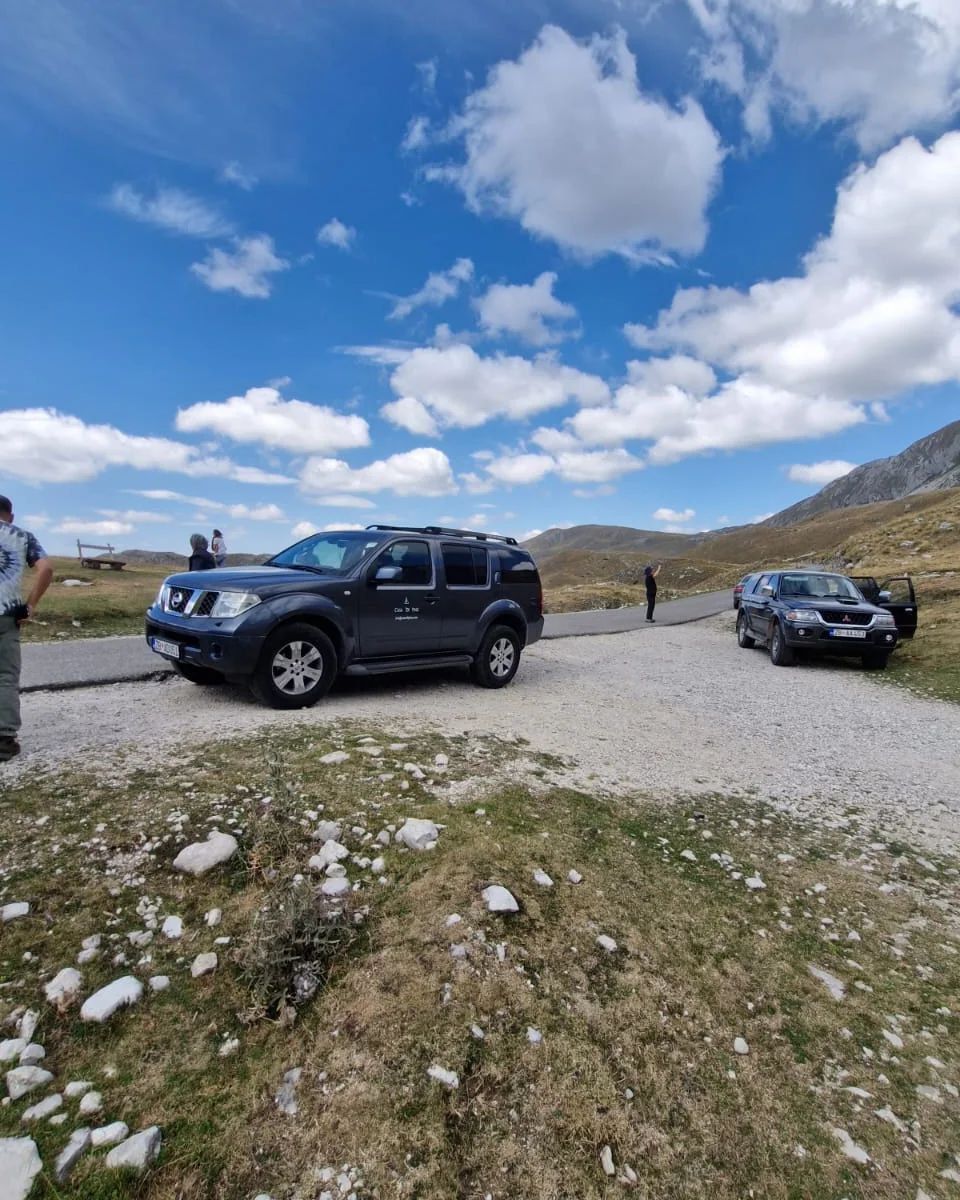 Two SUVs parked on a gravel road in a mountainous landscape under a blue sky with clouds.