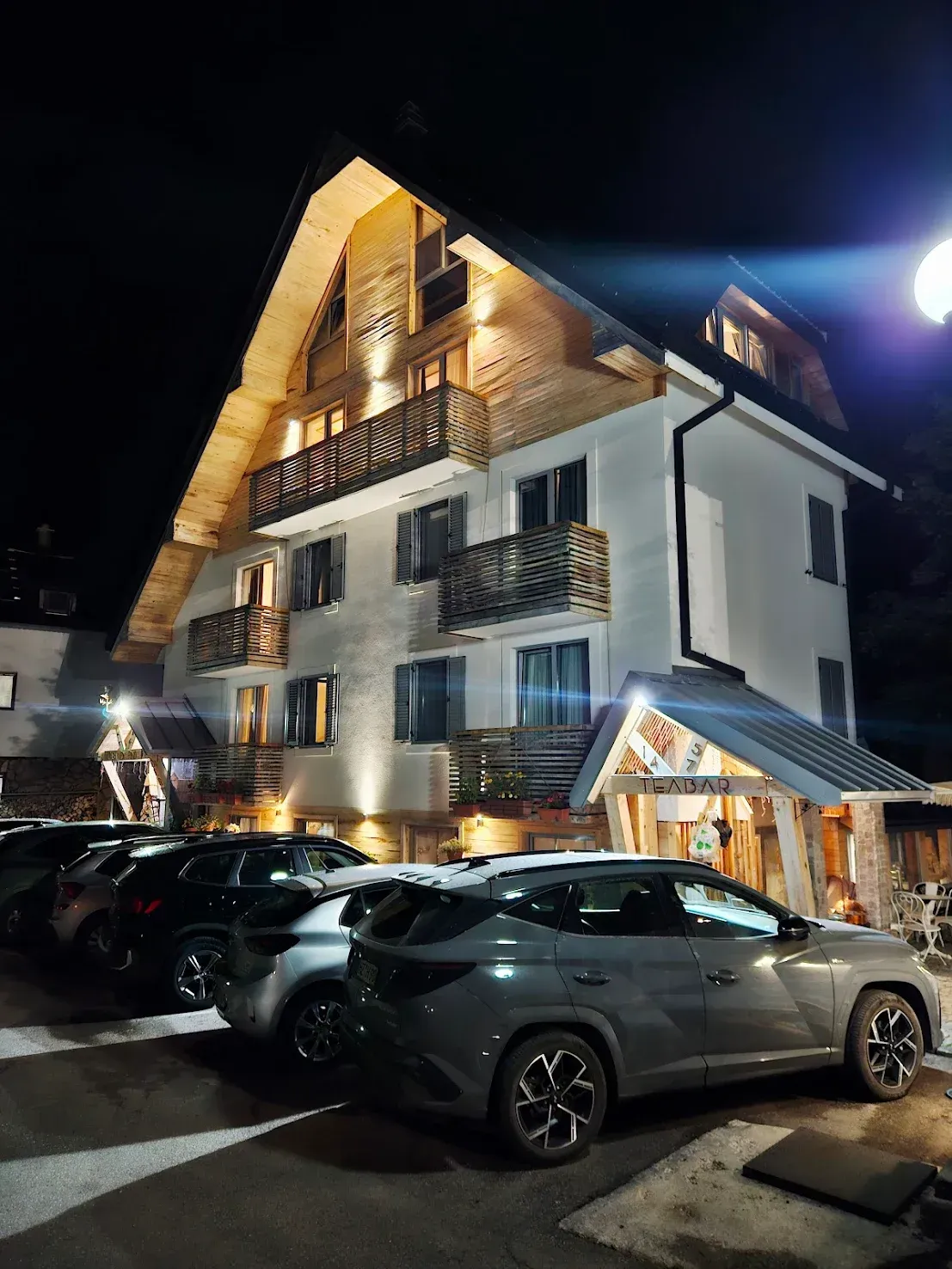 Hotel exterior at night with parked cars. Lit building, wooden accents, balconies.