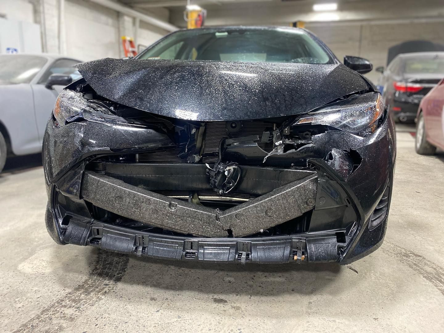 Black car with extensive front-end damage, likely from a collision, in an indoor garage setting.