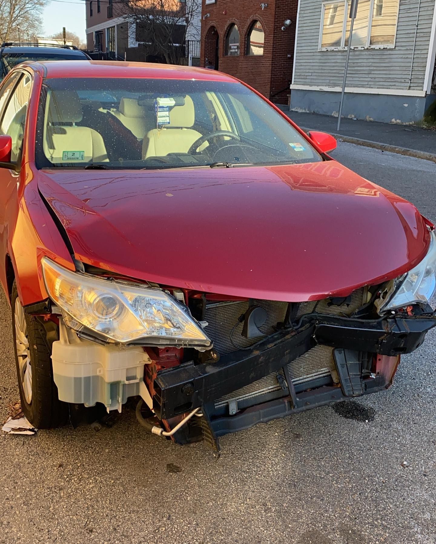 Red car with front-end damage parked on a street. The hood is crumpled, and parts are missing.
