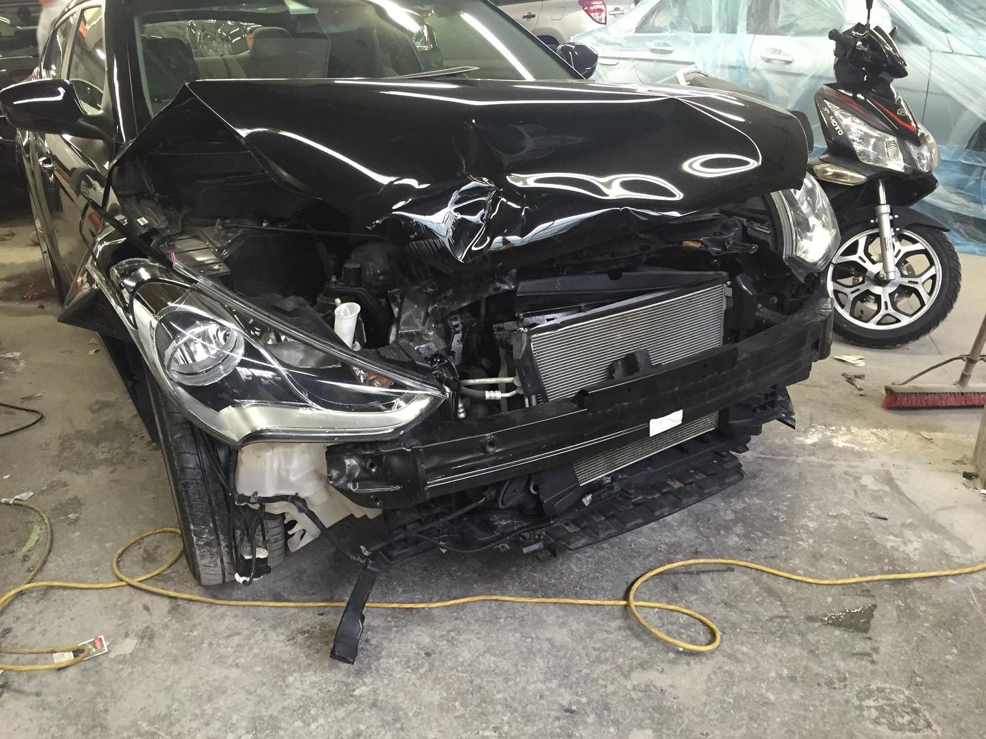 Damaged black car with extensive front-end collision damage in a repair shop, radiator exposed, next to a motorcycle.