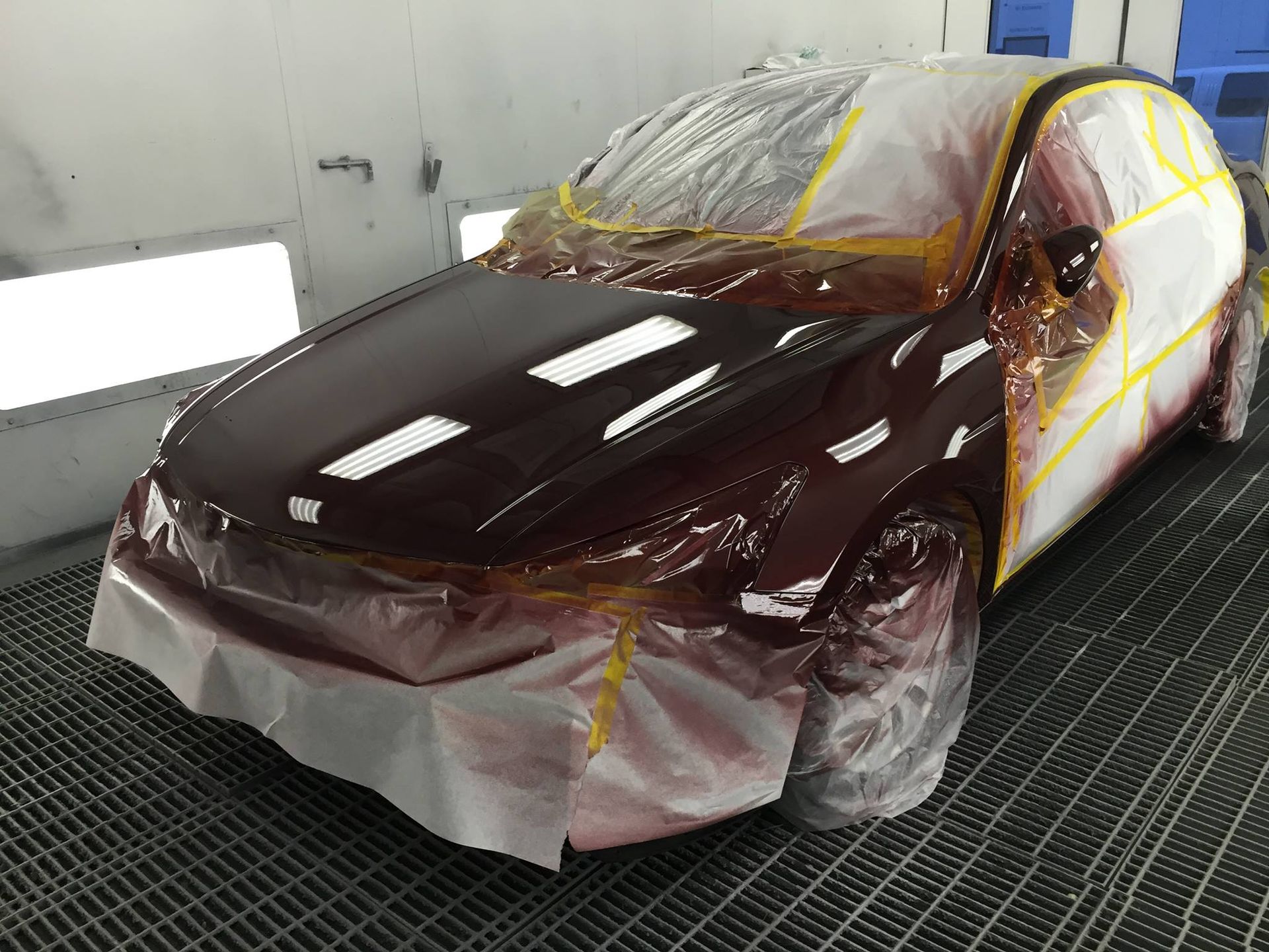 Car in a paint booth, partially masked with tape and plastic. Burgundy paint covers front.