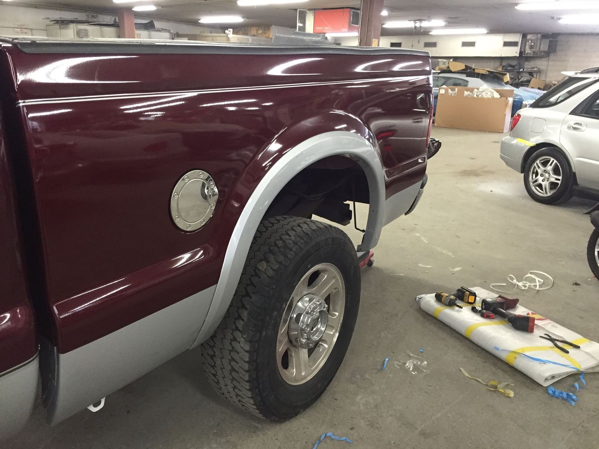 Burgundy and silver pickup truck in a shop, rear wheel exposed, tools on the floor.
