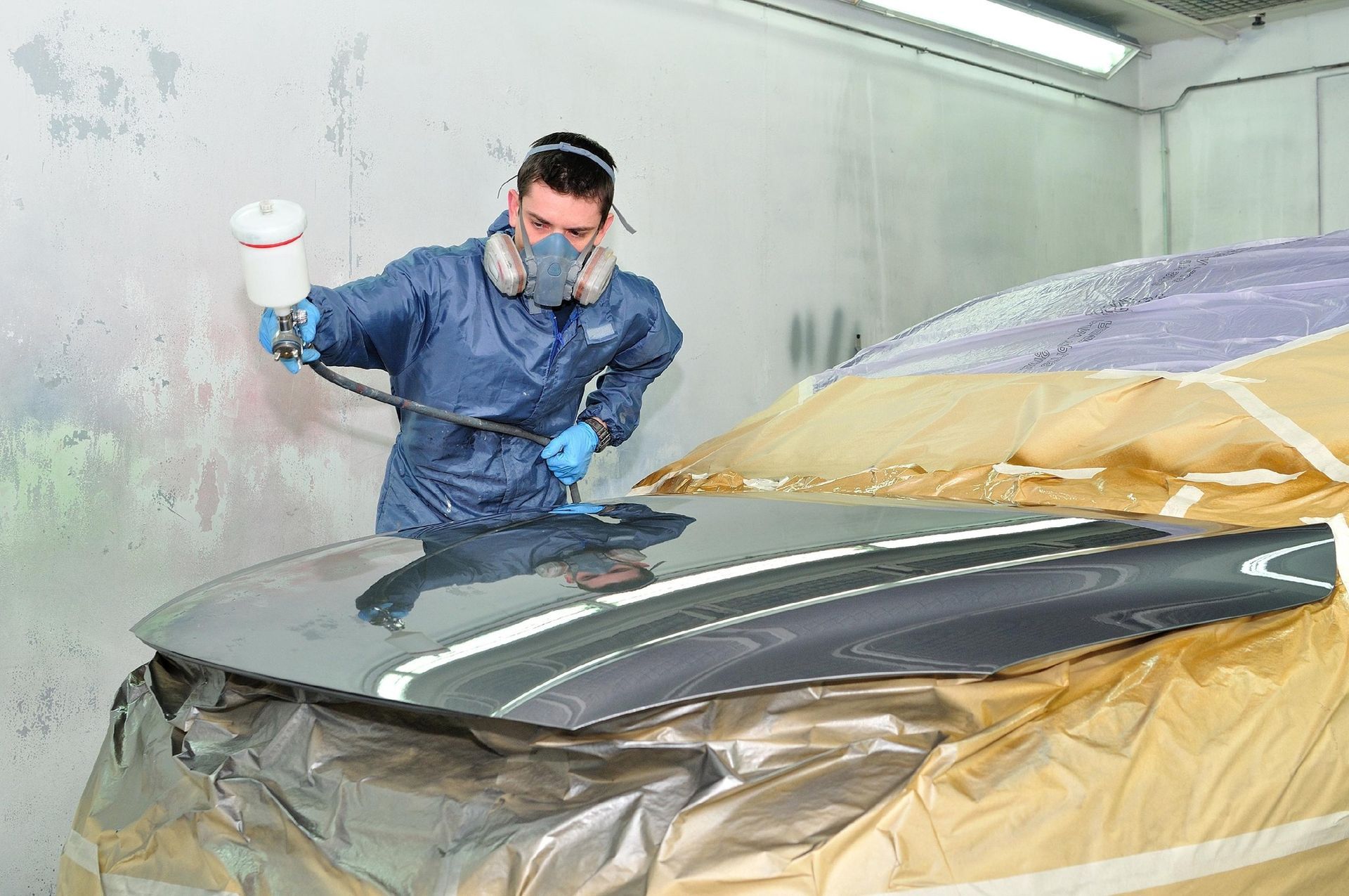 Person in protective suit spraying paint on car hood in a workshop.