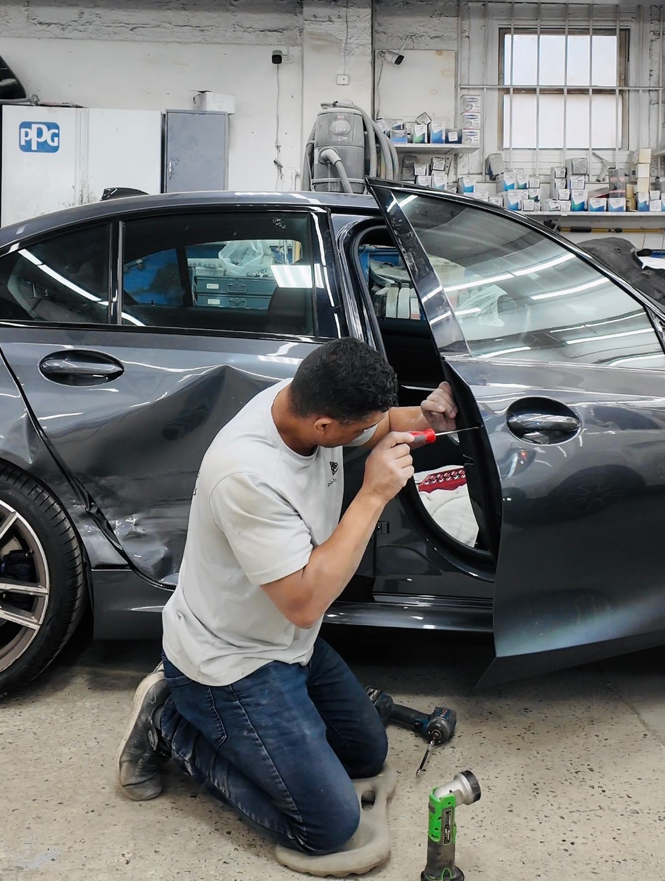Mechanic repairing a damaged gray car in a shop. He’s kneeling, working on the door with tools.