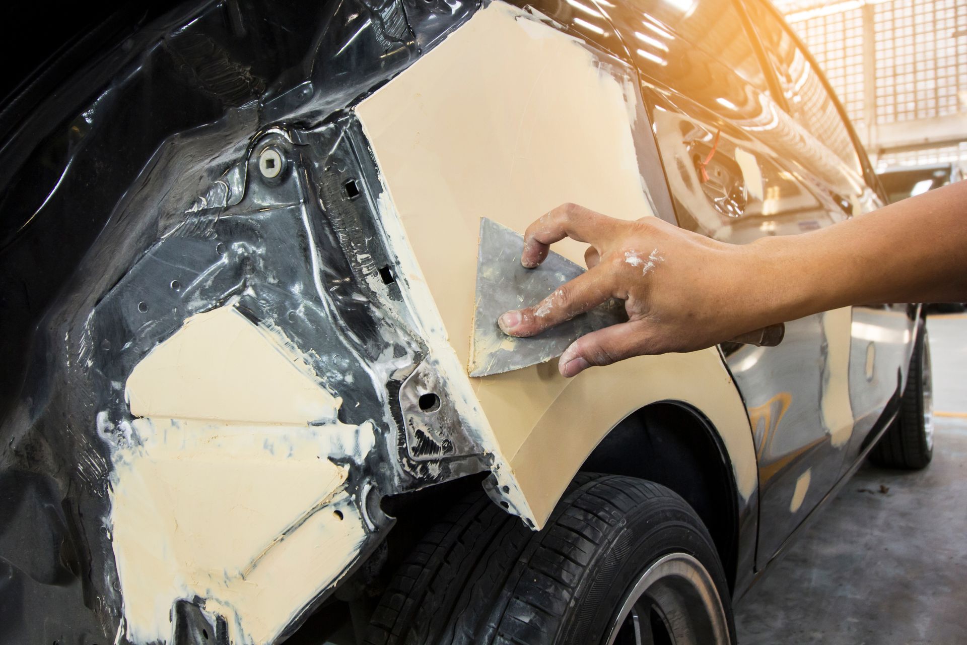 A person sanding body filler on a damaged car panel, in a repair shop.
