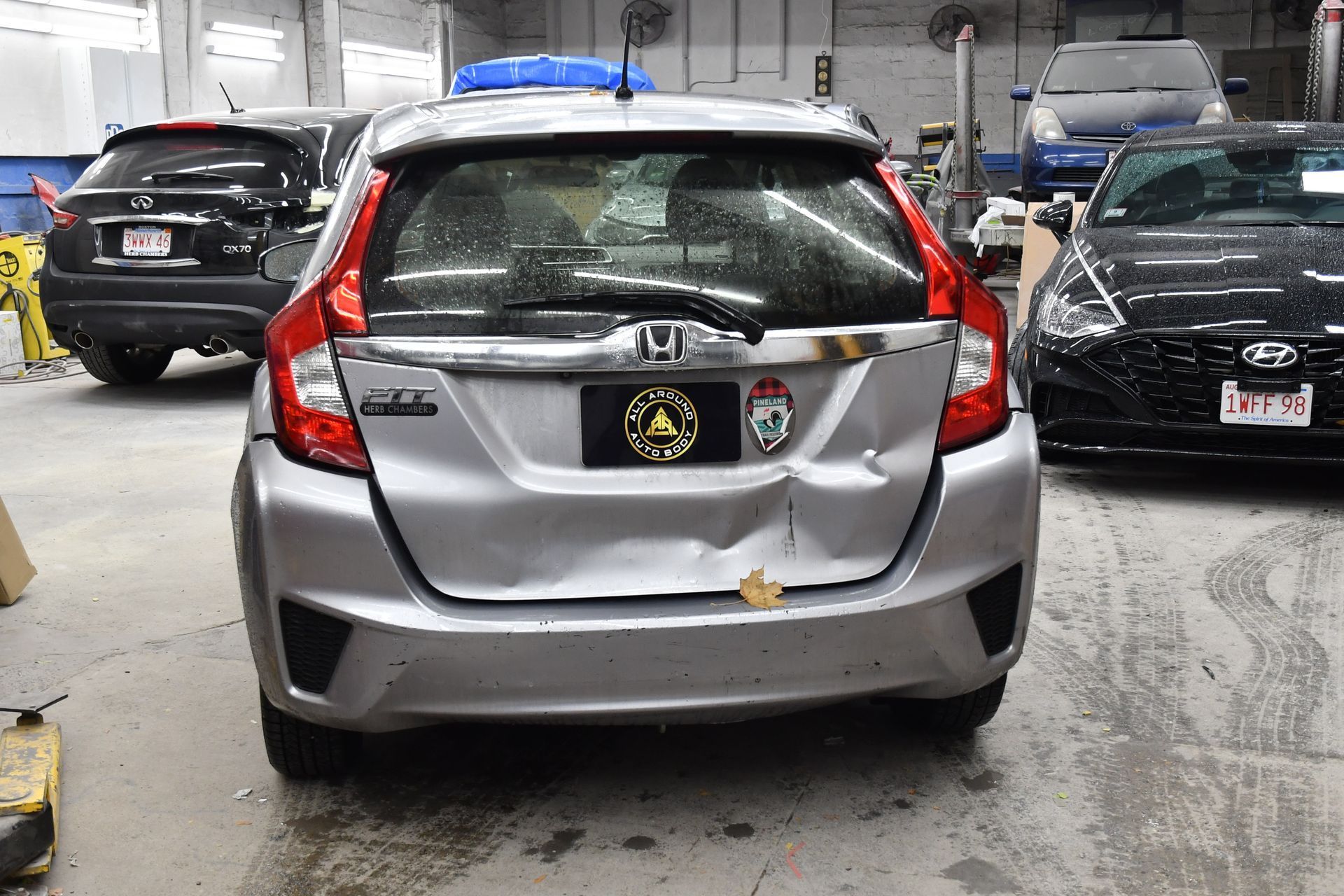 Silver Honda Fit with rear damage parked inside a garage, surrounded by other vehicles.