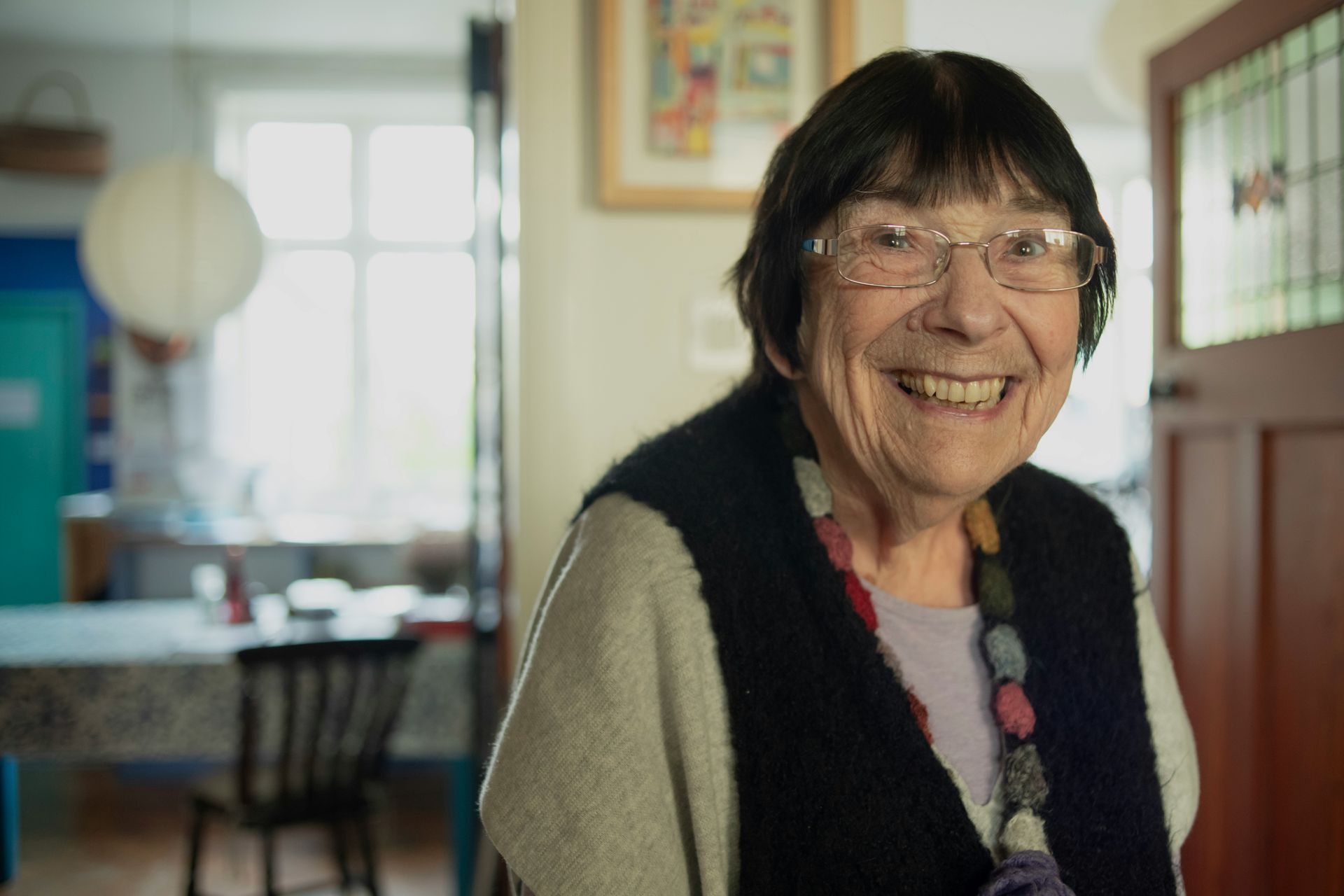 An elderly woman wearing glasses and a vest is smiling for the camera.